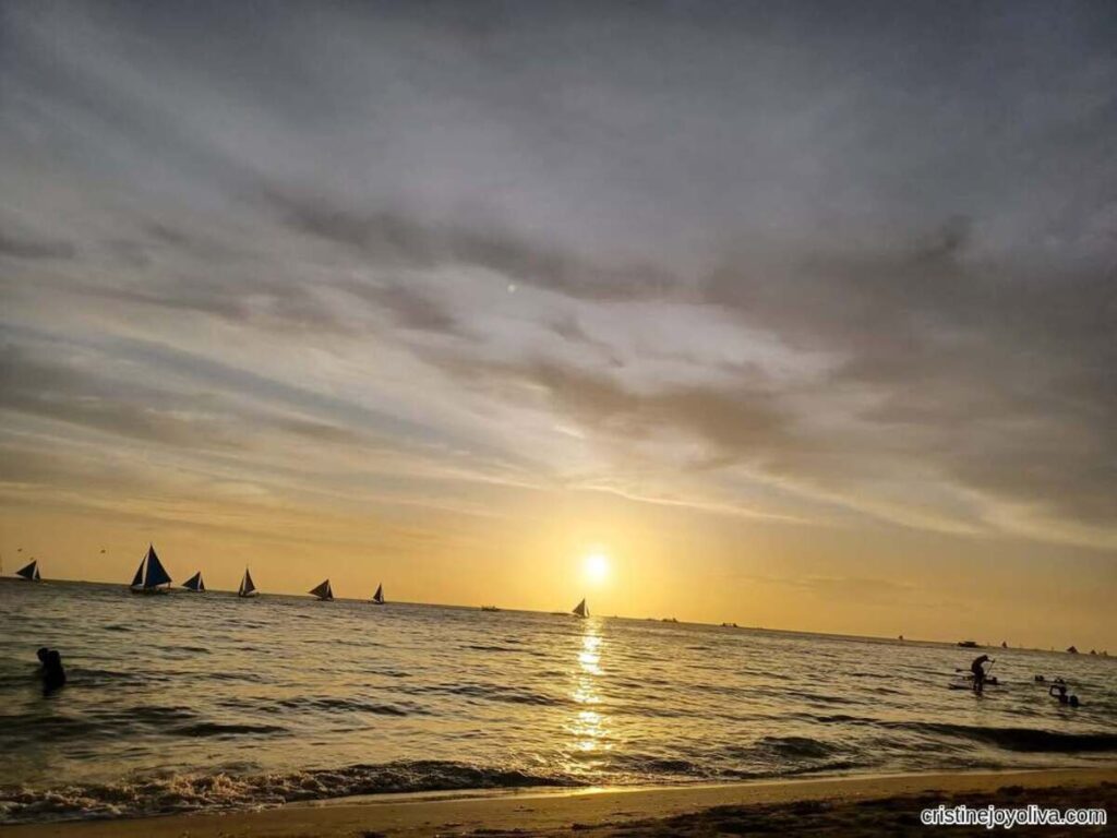 Boracay Beach at sunset with a golden sky, sailboats silhouetted on the ocean, people wading near the shore, and a sandy beach in the foreground under textured clouds.