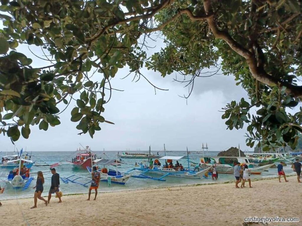 Colorful traditional boats with outriggers anchored near Boracay’s sandy shore, people walking along the beach, and leafy tree branches framing the calm ocean under cloudy skies.