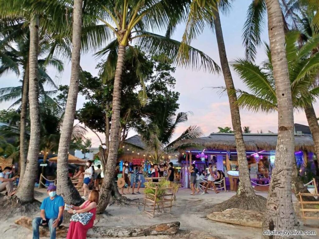 Beachside bar in Boracay with palm trees, bamboo seating, colorful lights, and people relaxing under a warm sunset sky.