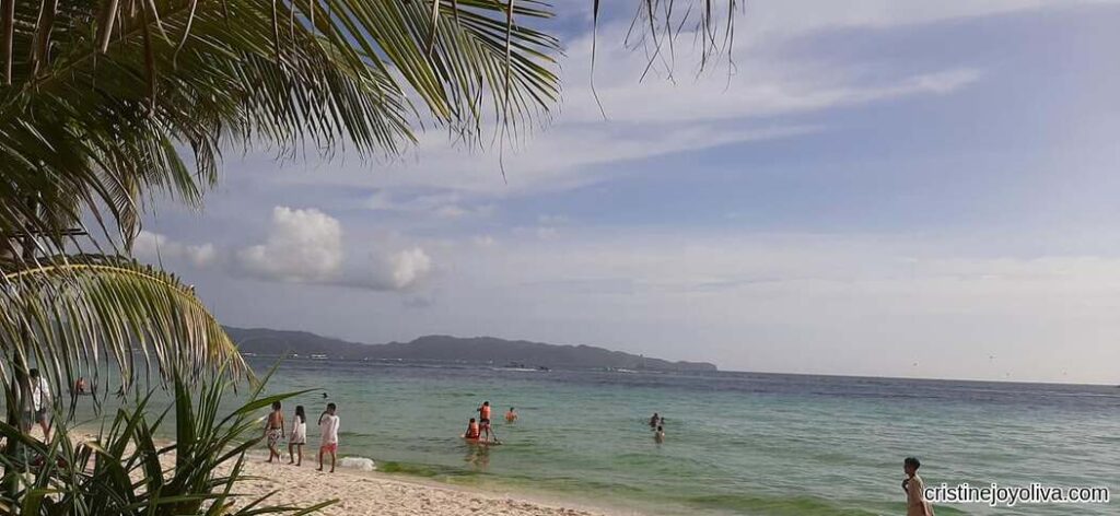 Boracay Beach with clear turquoise water, white sand, palm trees framing the view, and people walking and swimming near the shore under partly cloudy skies.