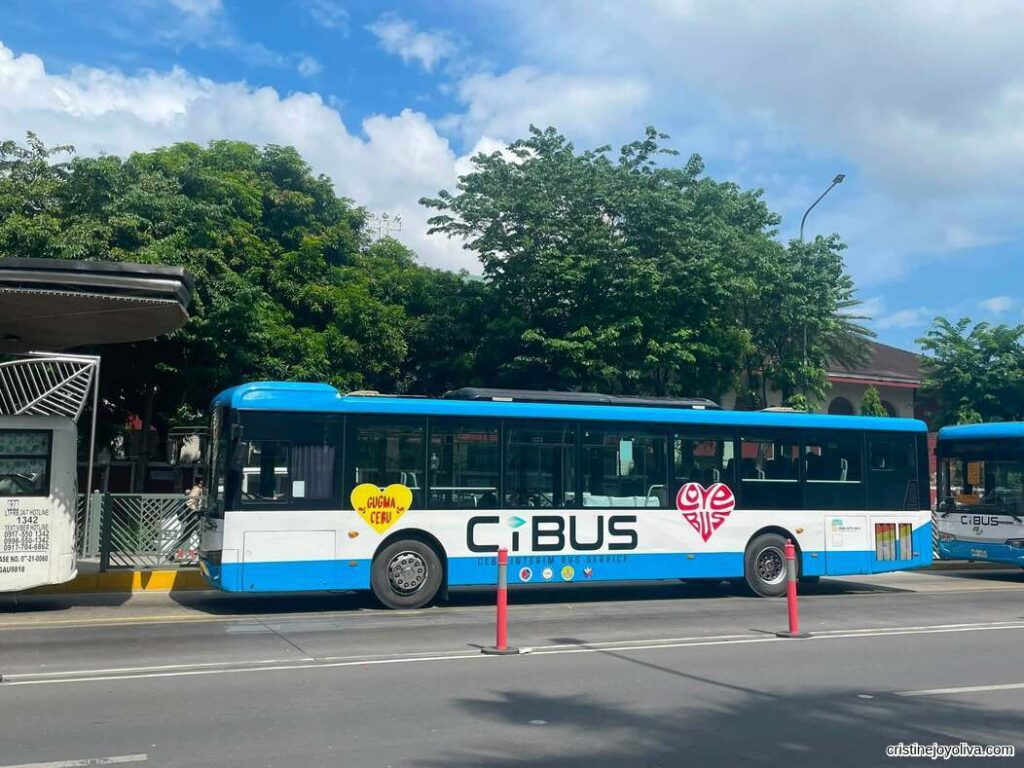 A blue and white CiBUS public transport bus parked at a terminal with trees and partly cloudy sky in the background