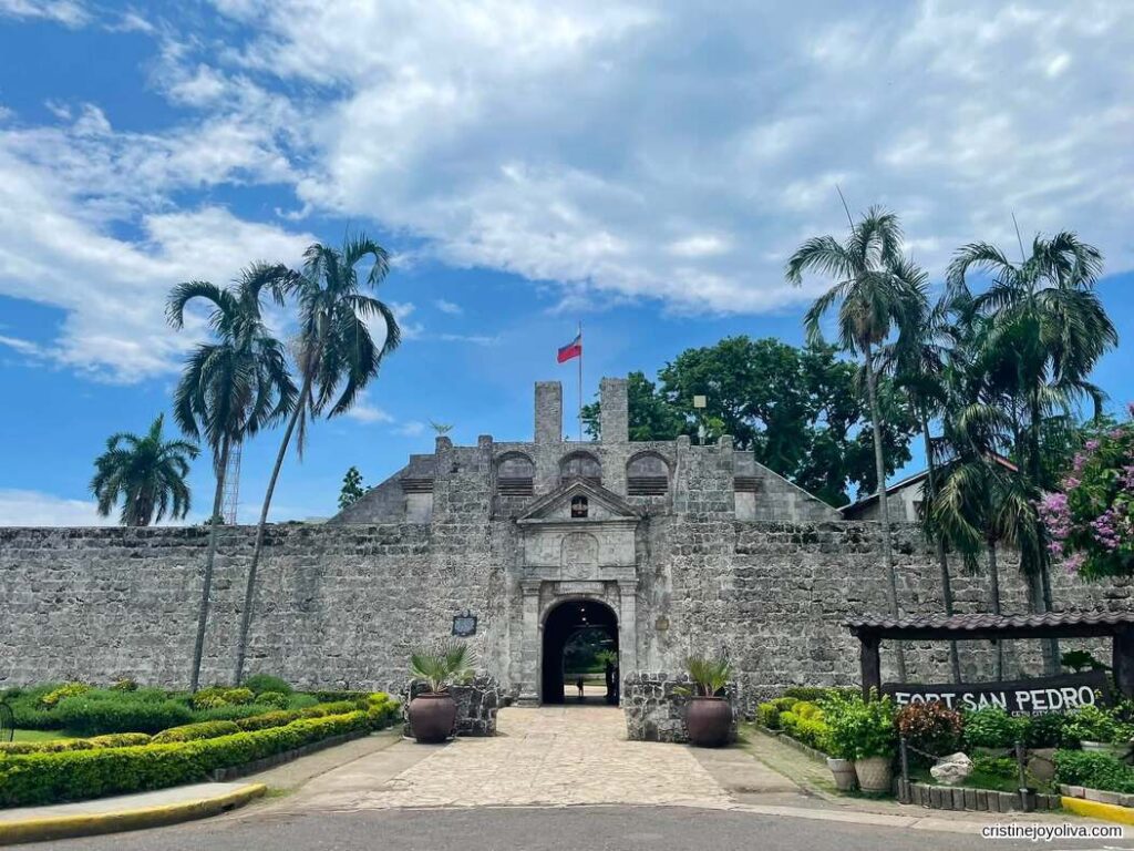 Entrance of Fort San Pedro, a historic stone fortress with palm trees and a Philippine flag under a partly cloudy sky