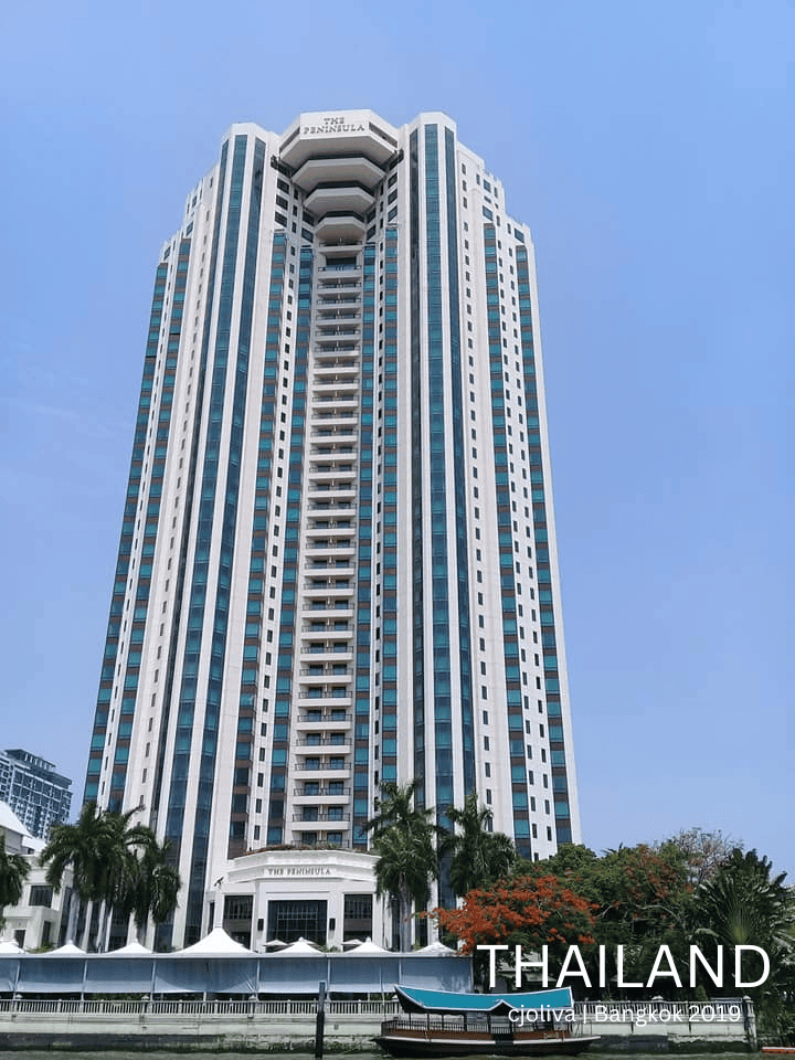 A tall white and blue high-rise building of The Peninsula Hotel with palm trees and river in foreground, Icon Siam, Bangkok