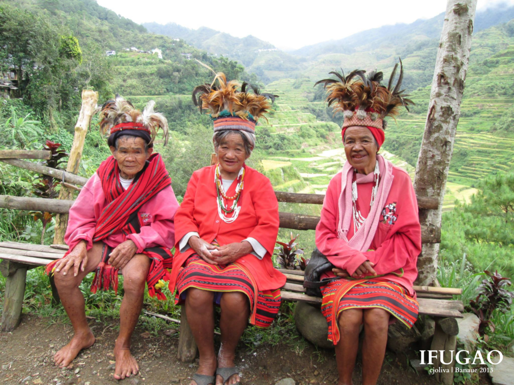 Three Ifugao elders wearing traditional woven red attire and feathered headdresses are seated on a wooden bench overlooking rice terraces.