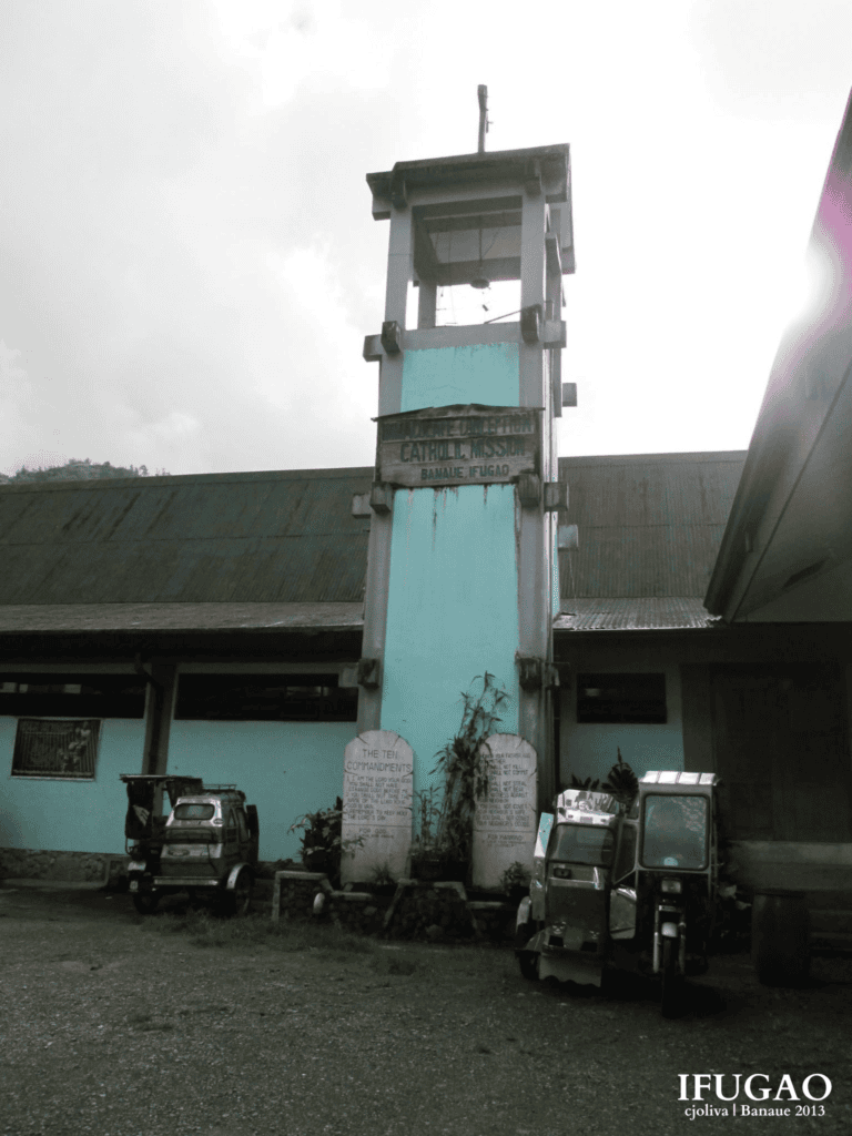 Bell tower and wooden sign of the Immaculate Conception Catholic Mission with parked tricycles in Banaue, Ifugao.