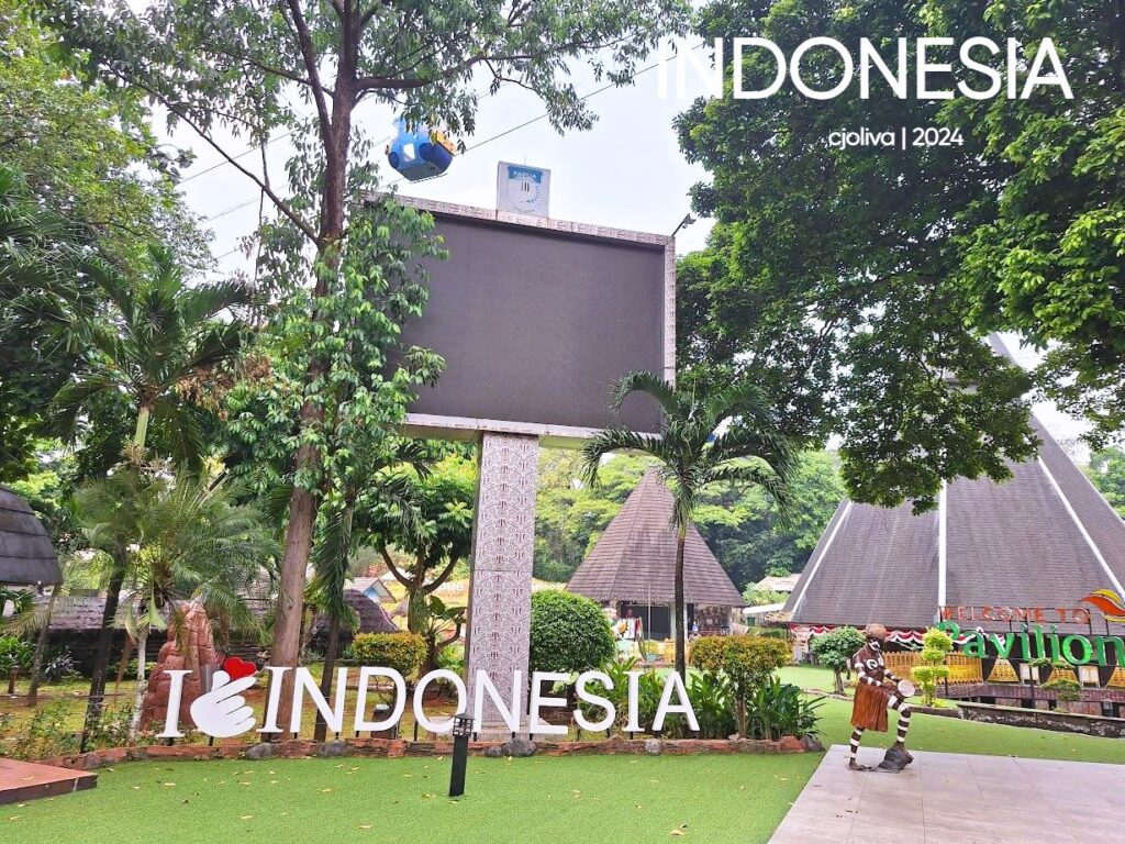Outdoor view of the Indonesia Pavilion with the “I ❤️ Indonesia” sign, lush greenery, traditional-style buildings with triangular roofs, a decorative billboard, and a gondola above the site.