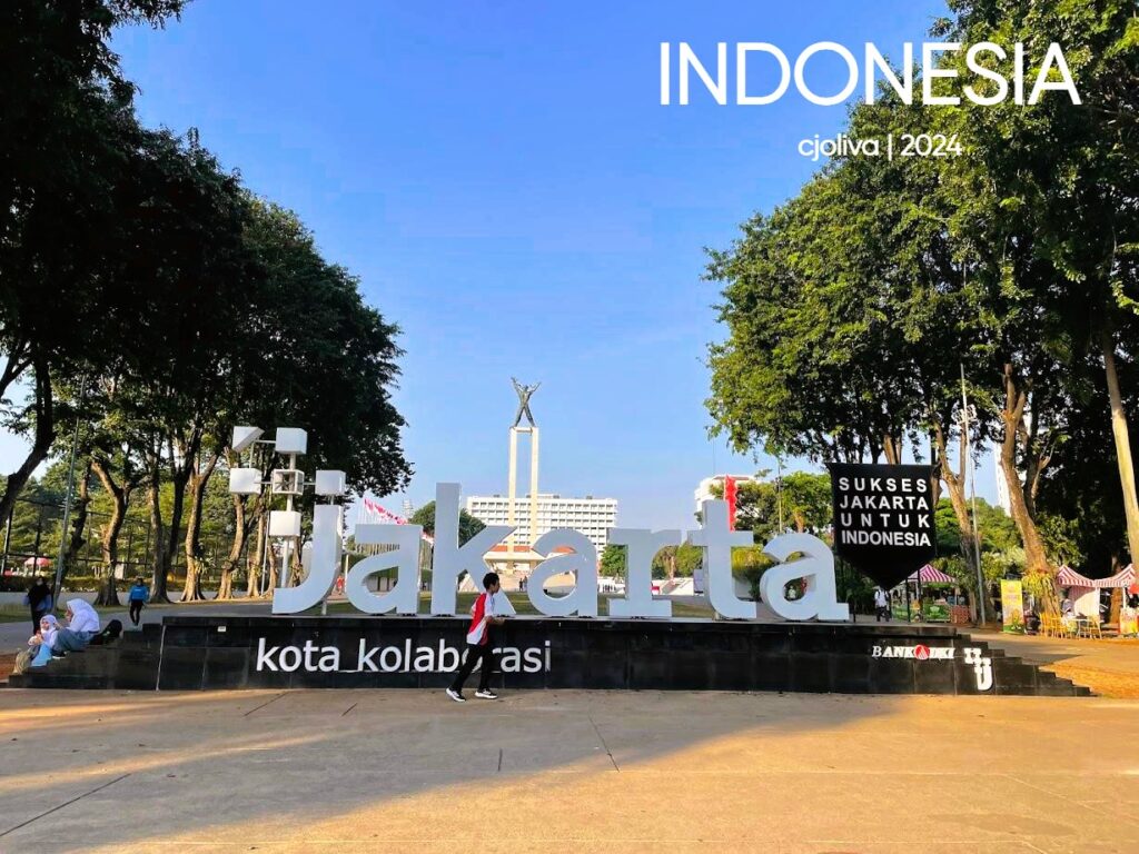 Public area in Jakarta featuring a large “Jakarta Kota Kolaborasi" sign, a nearby panel reading “Sukses Jakarta untuk Indonesia,” with a tall monument, trees, and city buildings in the background.