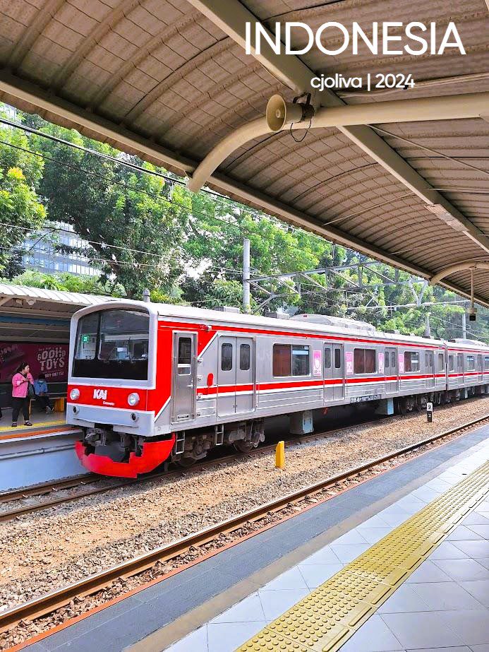 A KAI commuter train in Indonesia with a silver body and red-white stripes at a station platform, featuring a covered roof, tactile paving, and passengers waiting nearby.