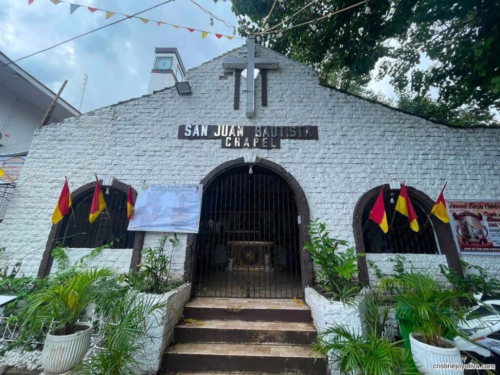 Front view of San Juan Bautista Chapel with white stone facade, arched windows, and entrance decorated with red and yellow flags