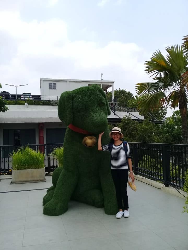A woman posing with large green dog-shaped topiary sculpture wearing a red collar with a bell at Taj Maharaj, Bangkok