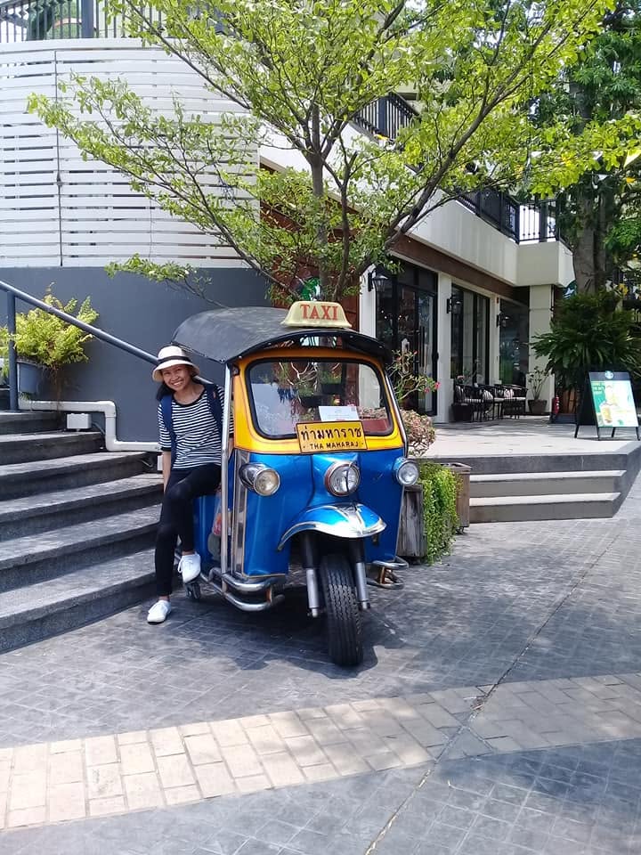 Smiling woman sitting on a blue tuk-tuk taxi with "Tha Maharaj" sign parked by steps and greenery in Bangkok