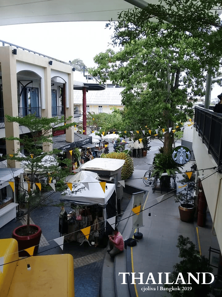 View of outdoor market stalls, greenery, and Starbucks sign at Taj Maharaj shopping area in Bangkok