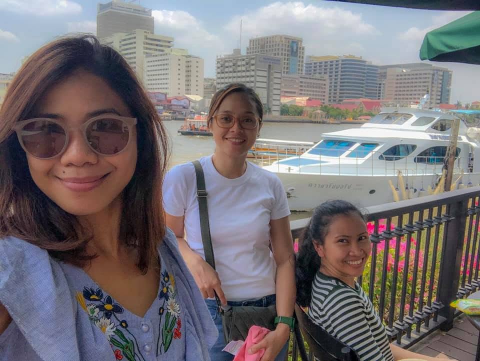Three smiling friends posing by the riverside with boats and city buildings in the background at Tha Maharaj, Bangkok