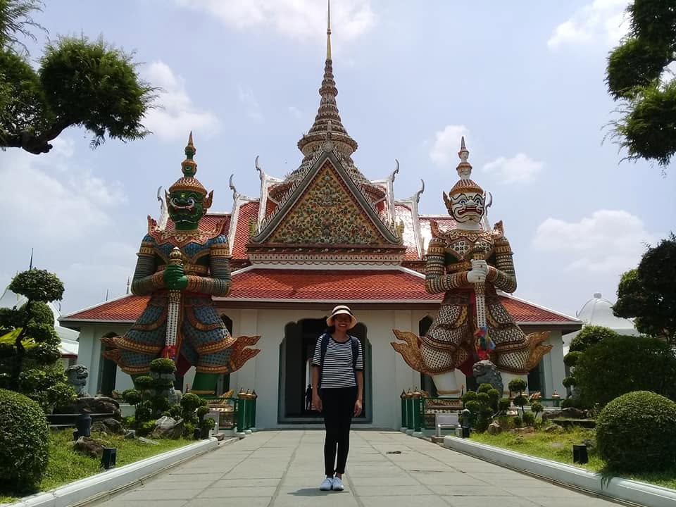 A woman standing between two large colorful guardian statues in front of ornate temple building at Wat Arun, Bangkok