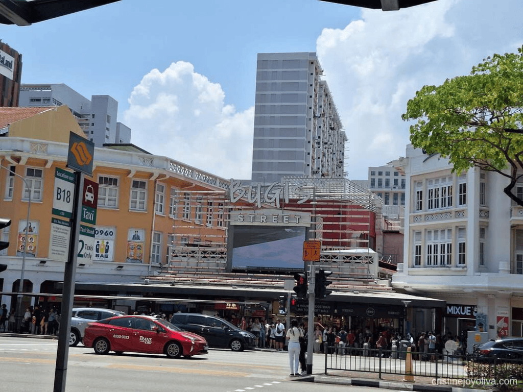 Street-level view of the vibrant Bugis Street entrance sign surrounded by historic colorful shophouses, modern high-rise apartments, and a red TransCab taxi on a sunny day in Singapore.