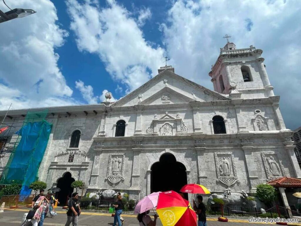 Cebu City solo travel: low-angle shot of the coral-stone facade and bell tower of the Basilica Minore del Santo Niño, with visitors holding colorful umbrellas under a blue sky.