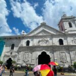 Cebu City solo travel: low-angle shot of the coral-stone facade and bell tower of the Basilica Minore del Santo Niño, with visitors holding colorful umbrellas under a blue sky.