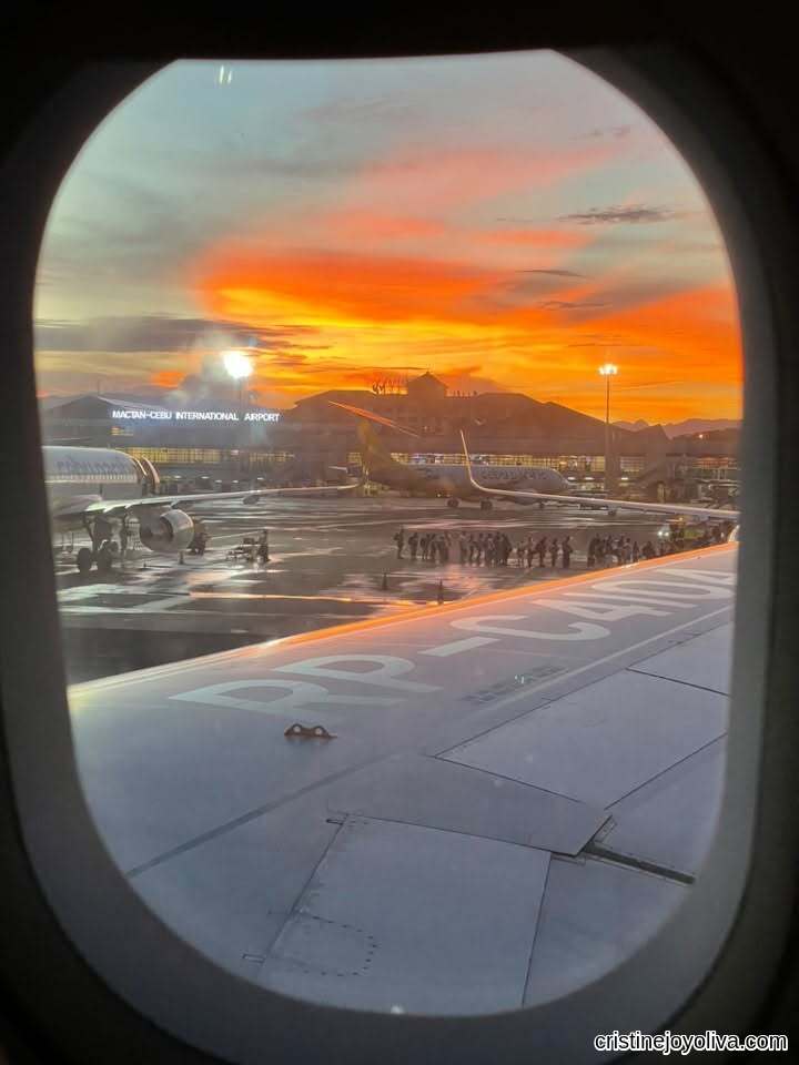 View through an airplane window showing an airplane wing on the tarmac at Mactan-Cebu International Airport against a bright fiery orange sunset, highlighting Cebu City solo travel.