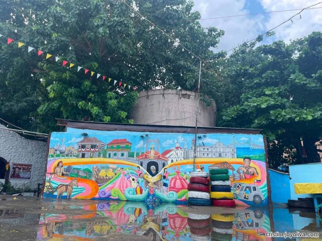 Wide shot of a bright outdoor mural wall under lush green trees featuring colorful paintings of Cebu landmarks, churches, a bridge, and Sinulog dancers, with painted tires stacked in front and reflections casting on wet ground.