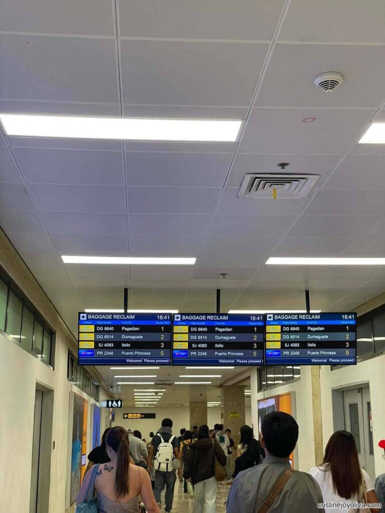 Passengers walking down an airport terminal hallway toward three digital overhead baggage reclaim screens displaying flight numbers for Pagadian, Dumaguete, Iloilo, and Puerto Princesa.