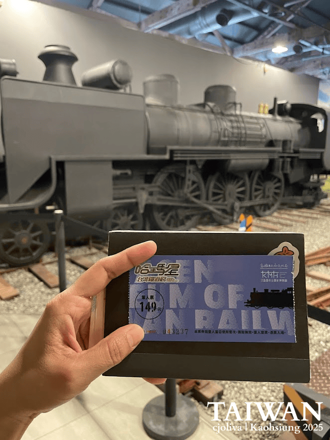 A hand holding an admission ticket in front of a vintage black steam locomotive inside the Hamasen Museum of Taiwan Railway in Kaohsiung.