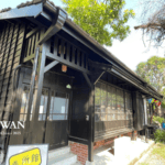 Traditional wooden building at Hinoki Village in Chiayi, Taiwan, with dark wood paneling, a tiled roof, colorful lanterns, landscaped greenery, and visitors exploring the cultural site.