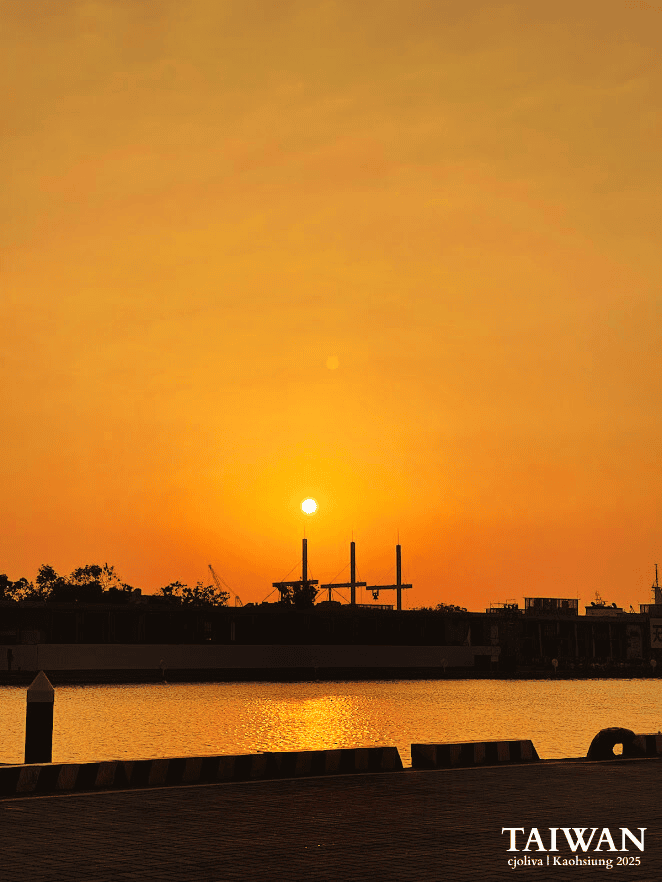 Silhouetted harbor cranes and industrial structures along the waterfront as the sun sets over warm golden waters in Kaohsiung, Taiwan.