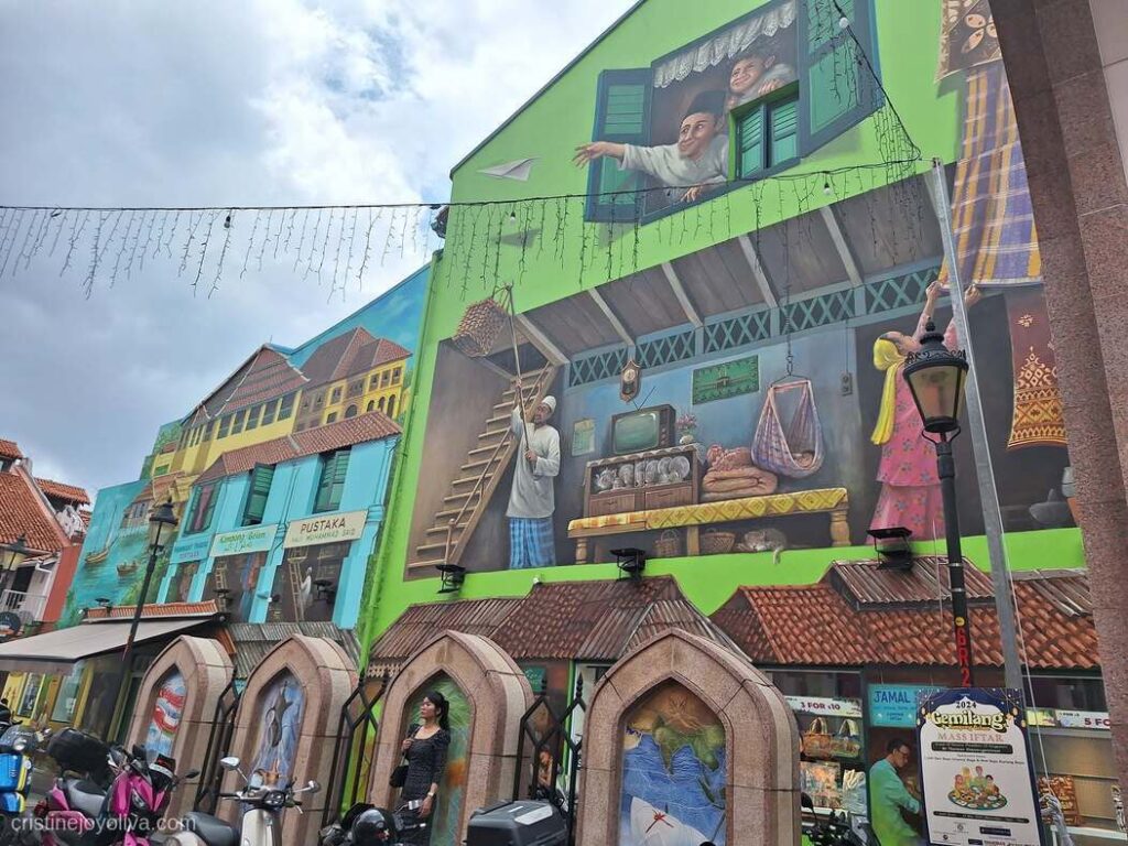 Large heritage mural on a green building wall in Kampong Glam, Singapore, depicting traditional Malay life, local merchants, and shophouse architecture along Muscat Street.