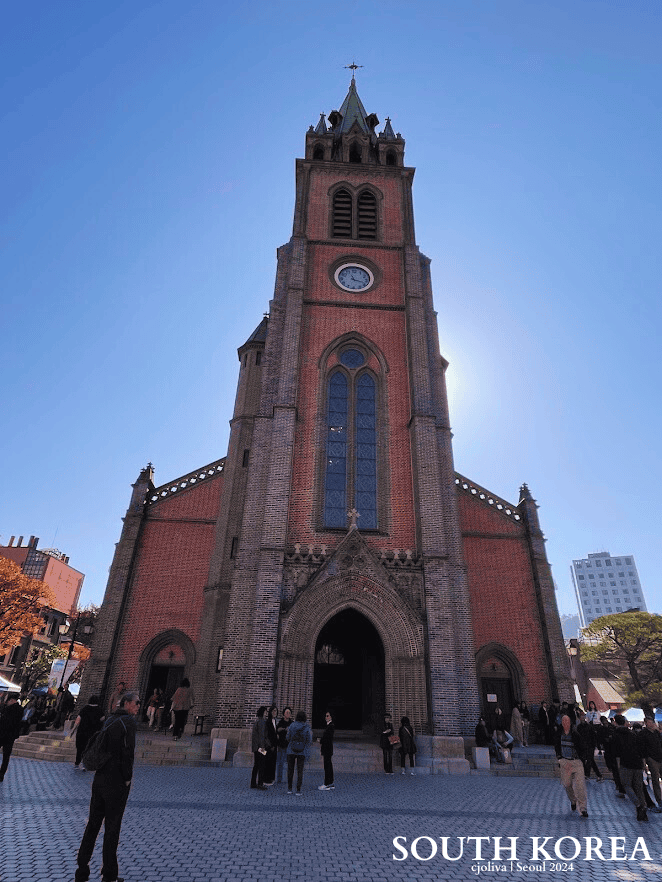 A low-angle view of the historic Myeongdong Cathedral in Seoul, South Korea. The red brick Gothic Revival church features a tall clock tower and central spire against a bright blue sky with people gathered at the entrance.
