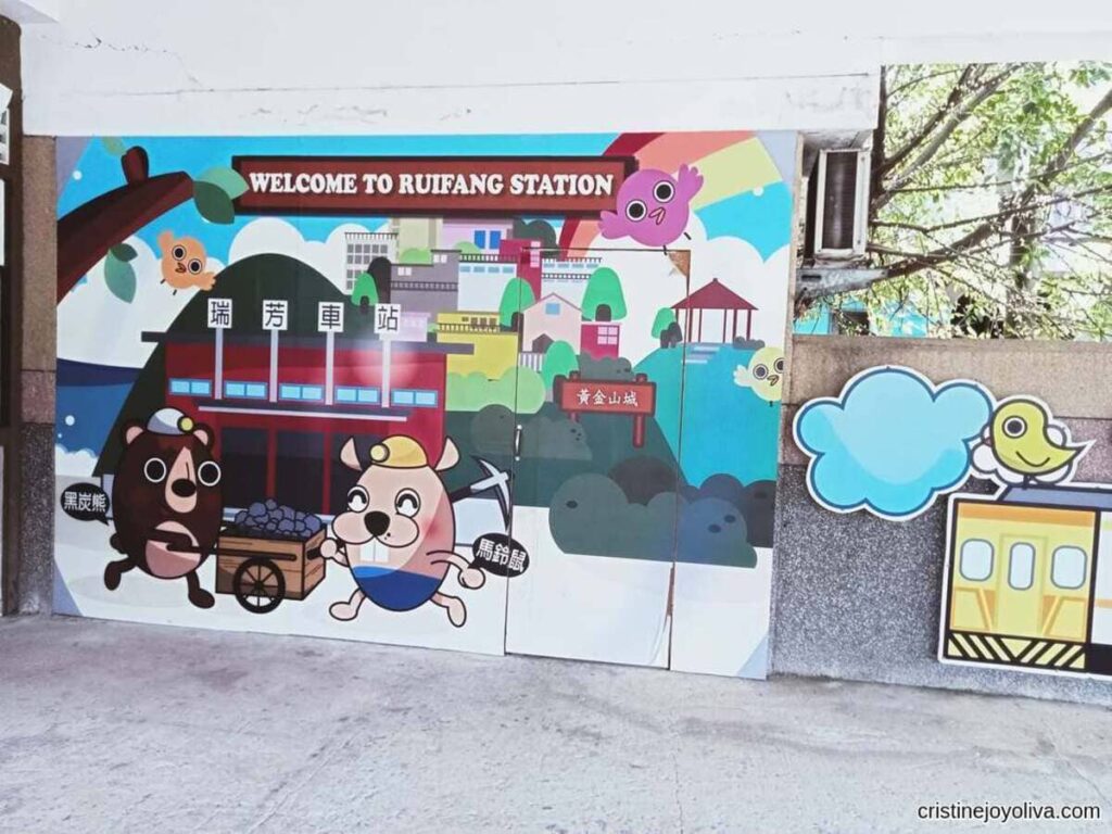 A "Welcome to Ruifang Station" mural featuring cartoon coal-mining mascots pushing a cart, with birds and a village backdrop.