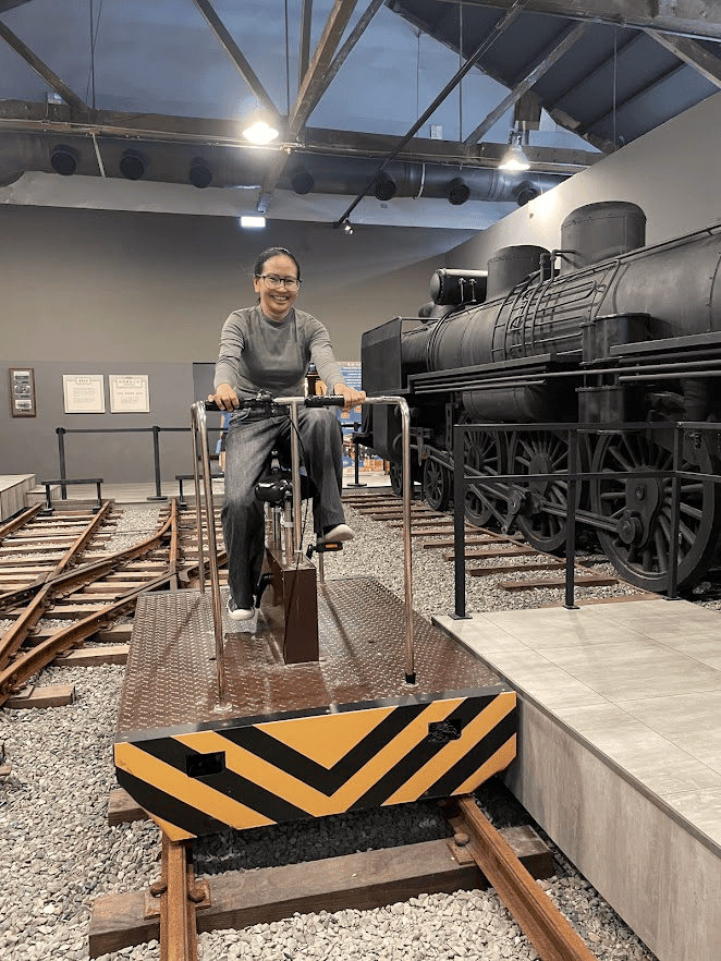 A smiling woman riding a handcar rail bike exhibit mounted on track rails next to a vintage black steam train at the Hamasen Museum of Taiwan Railway.
