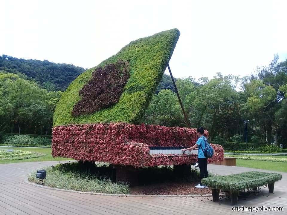 A large grand piano sculpture covered in green and red plants with a person playing the keys, located at Shilin Residence Park in Taipei, Taiwan.