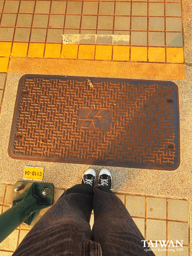 Top-down view of a person wearing black canvas sneakers and jeans standing on a paved sidewalk next to a textured metal utility cover in Kaohsiung, Taiwan.
