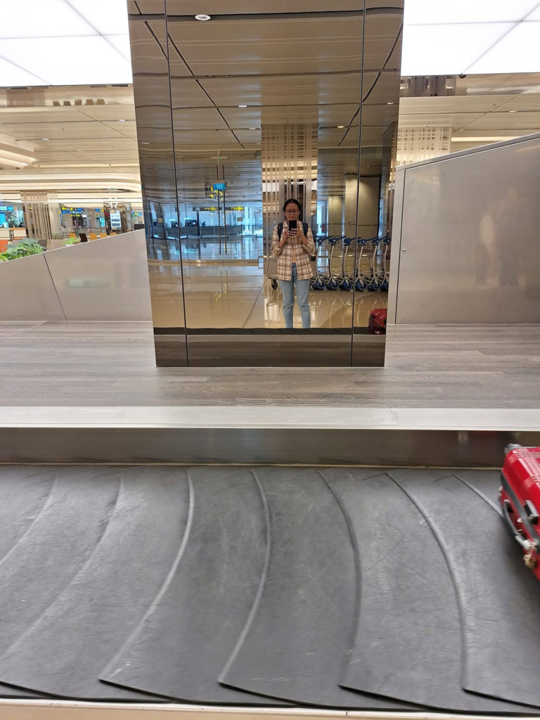 Solo female traveler mirror selfie at Changi Airport Terminal 1 baggage claim carousel. A traveler in a plaid shirt and jeans reflects in a polished pillar while waiting for luggage.