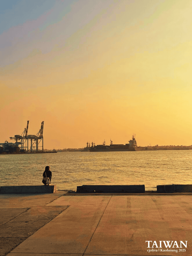 Silhouette of a solo traveler sitting on a pier watching the golden sunset over cargo ships at Kaohsiung Harbor in Taiwan.