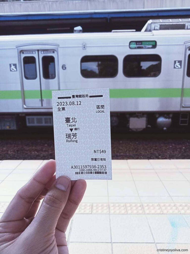 A hand holding a Taiwan Railway one-way ticket from Taipei to Ruifang, priced at NT$49, with a train in the background.