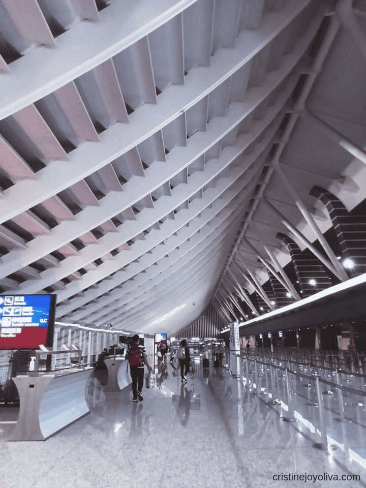 Interior of Taiwan Taoyuan International Airport terminal featuring rib-like curved ceiling beams, polished reflective floors, check-in counters, digital flight boards, and travelers with luggage.