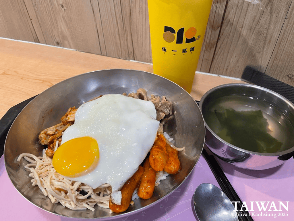 Taiwanese lunch in Kaohsiung, featuring a metal bowl with rice topped by a sunny-side-up egg, grilled chicken, noodles, and sausages, served with seaweed soup and a tall yellow drink cup.