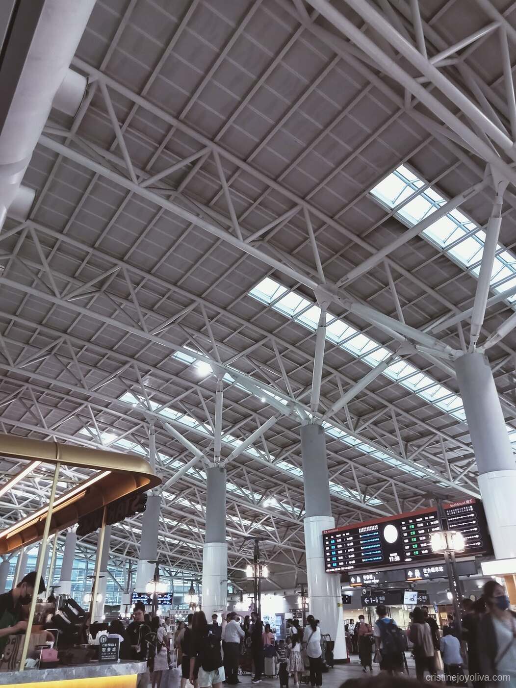 Interior of Zuoying High-Speed Rail Station in Kaohsiung, Taiwan, with steel beam ceiling, skylights, cylindrical columns, bilingual digital schedule board, travelers with luggage, and a coffee stand.