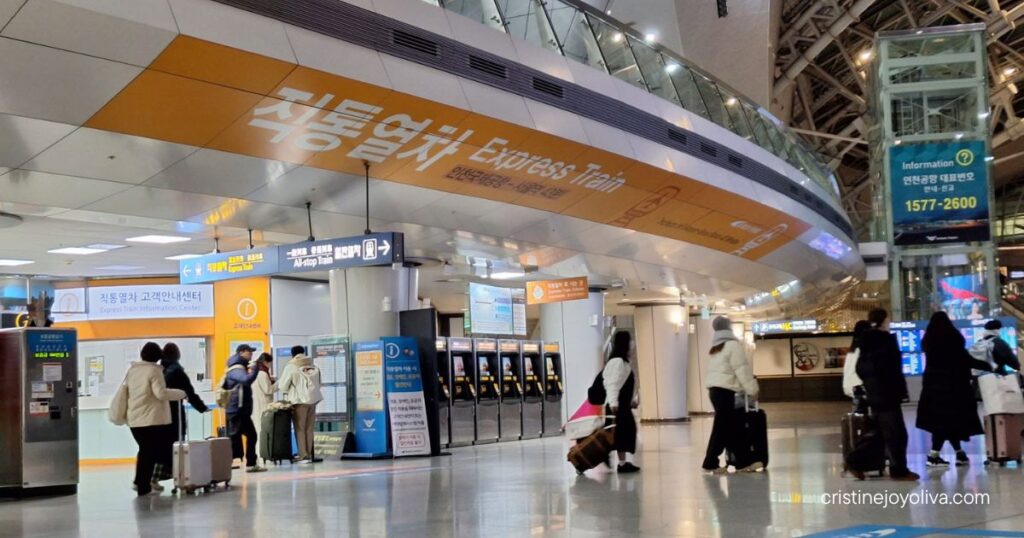 Travelers with luggage walk past AREX Express Train signs and ticket gates inside Incheon Airport.