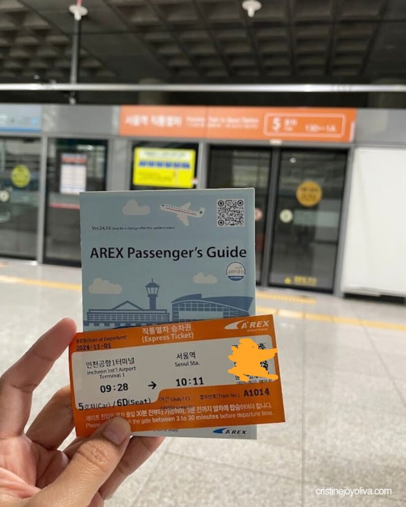 Hand holding an AREX Express ticket from Incheon Airport to Seoul Station in front of a train platform