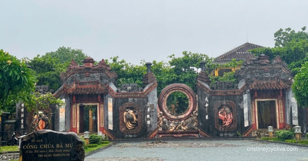 An ornate stone temple gate with intricate carvings and a circular center opening behind a stone sign at Ba Mu Temple in Hoi An.