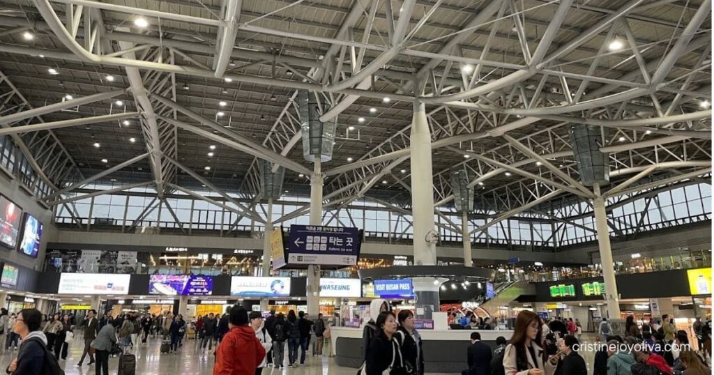 Busy Busan Station concourse with a white steel-truss ceiling, platform signs, and travelers with luggage