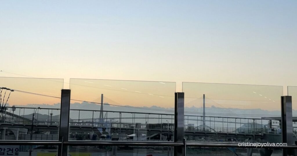 The sunrise sky is seen through glass panels at Busan Station, with a distant bridge and mountains in the background.