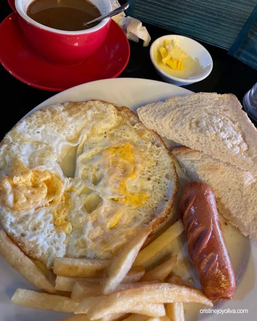 A white plate filled with fried eggs, French fries, a grilled sausage, and toast next to a red coffee cup.