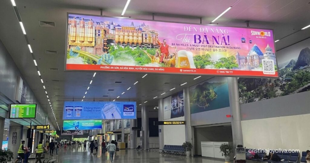 Passengers walk through a spacious, modern airport terminal underneath a brightly lit advertisement for Sun World Ba Na Hills in Da Nang.