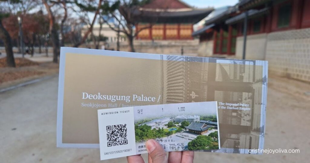 Hand holding a Deoksugung Palace admission ticket priced at 1,000 KRW, with the palace visible in the background