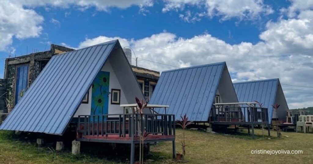 A row of blue metal-roofed A-frame glamping cabins with wooden front porches set on a grassy lawn under a blue sky with scattered clouds.