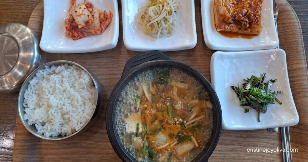 A bowl of hot Korean soybean paste stew sits on a wooden tray with steamed rice and small banchan side dishes.