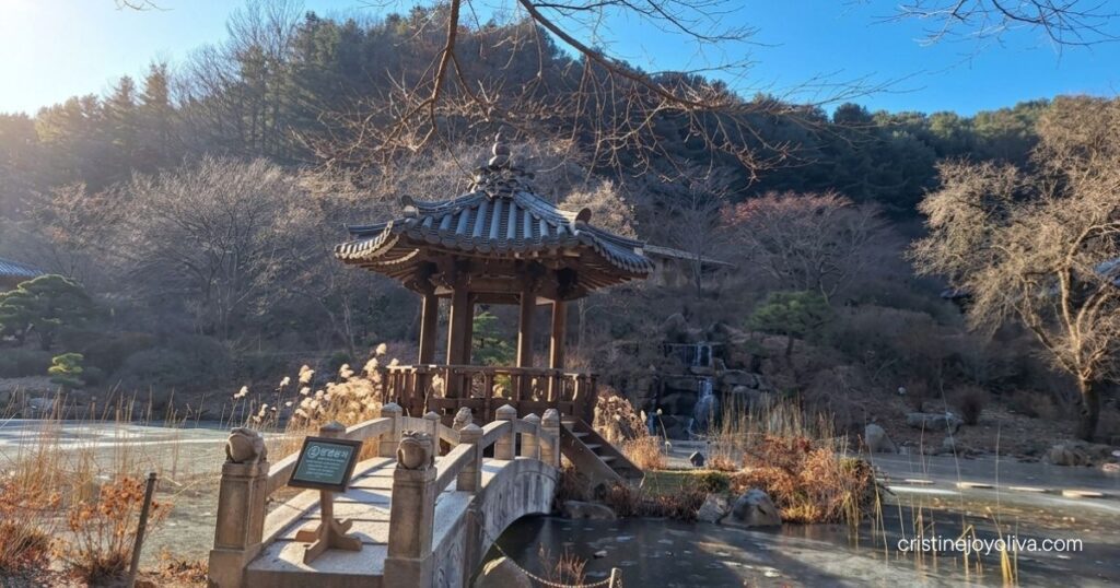 A stone footbridge leads to a traditional wooden pavilion beside a partially frozen pond at the Garden of Morning Calm.